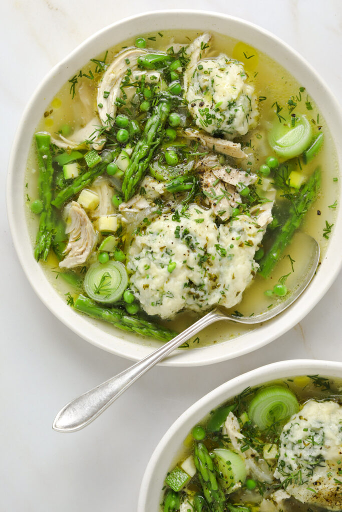 a white bowl with chicken and dumplings soup with asparagus, peas, zucchini, leeks and chopped herbs.