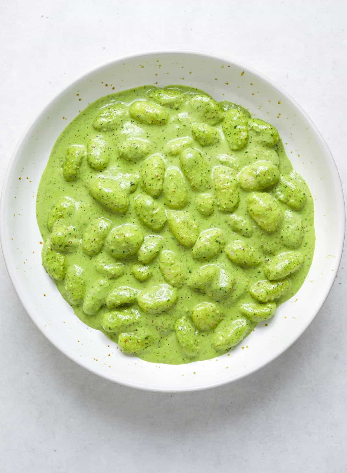 A white ceramic bowl of gnocchi with a green pasta sauce on a blue background.