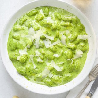 A bowl of green pesto gnocchi on a blue table with a bottle of wine, a glass of white, parmesan cheese and antique cutlery.
