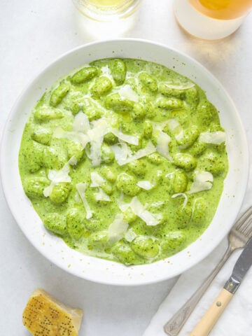 A bowl of green pesto gnocchi on a blue table with a bottle of wine, a glass of white, parmesan cheese and antique cutlery.