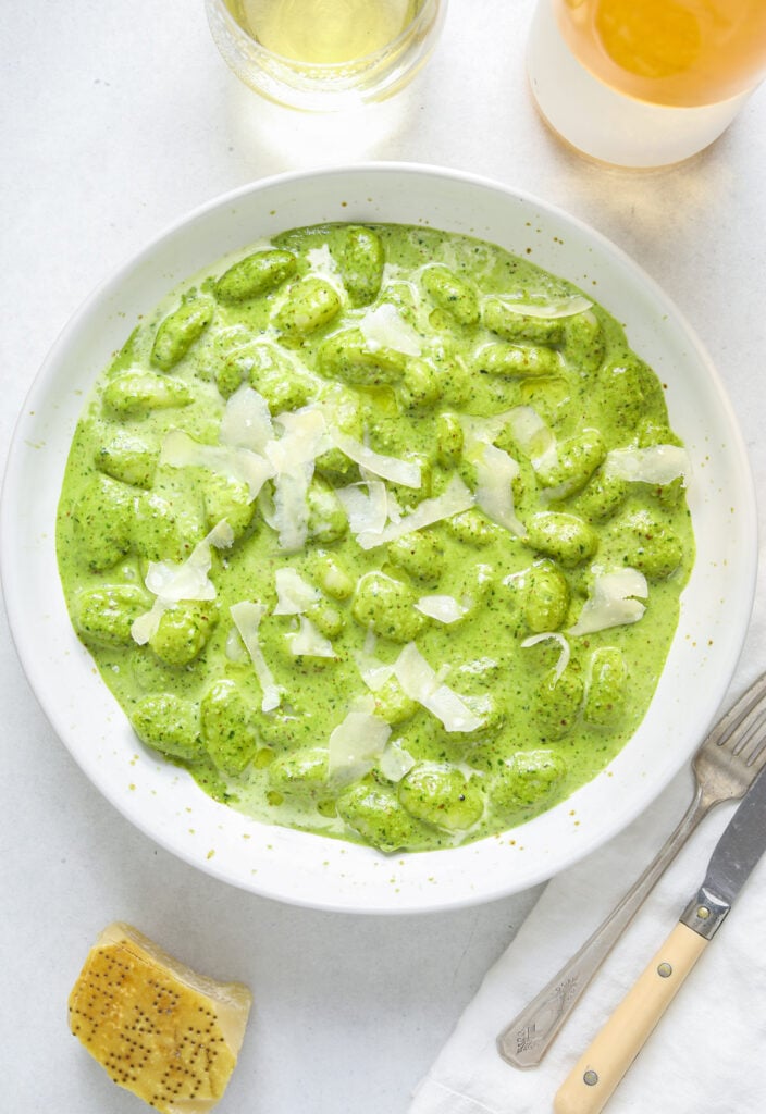 A bowl of green pesto gnocchi on a blue table with a bottle of wine, a glass of white, parmesan cheese and antique cutlery.