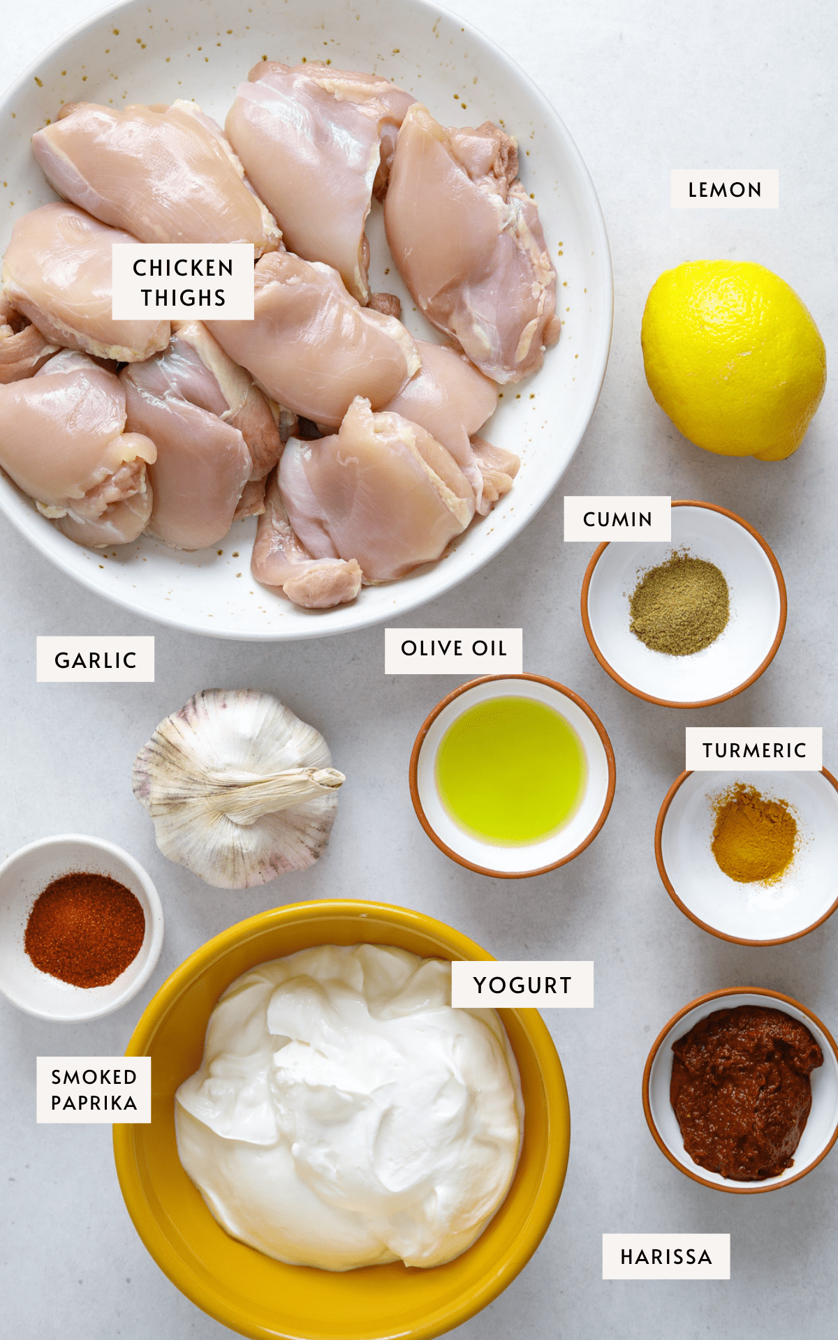 A bowl of raw chicken thighs, lemon, a bulb of garlic, a bowl of greek yogurt and spices in small bowls.