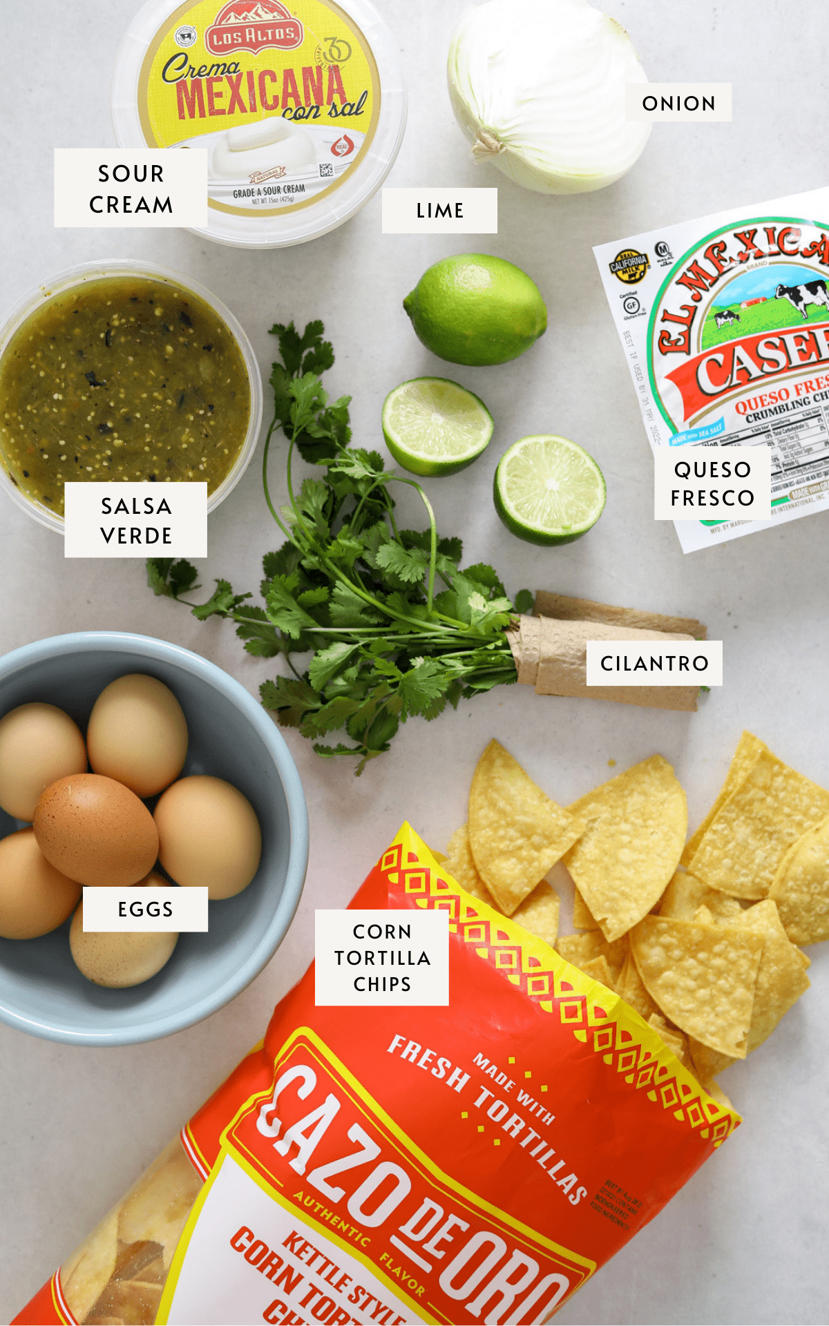 a container of tomatillo salsa, sour cream, queso fresco, a red bag of corn tortilla chips, a bundle of cilantro, a blue bowl filled with brown eggs, two limes and a white onion.