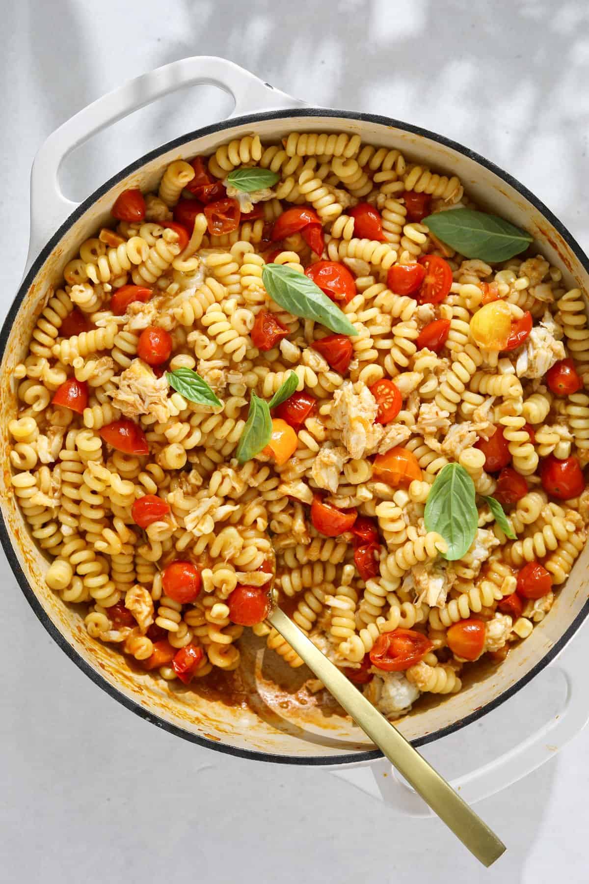 An enamel dutch oven pan filled with pasta with crab, tomatoes and fresh basil leaves with a gold spoon.