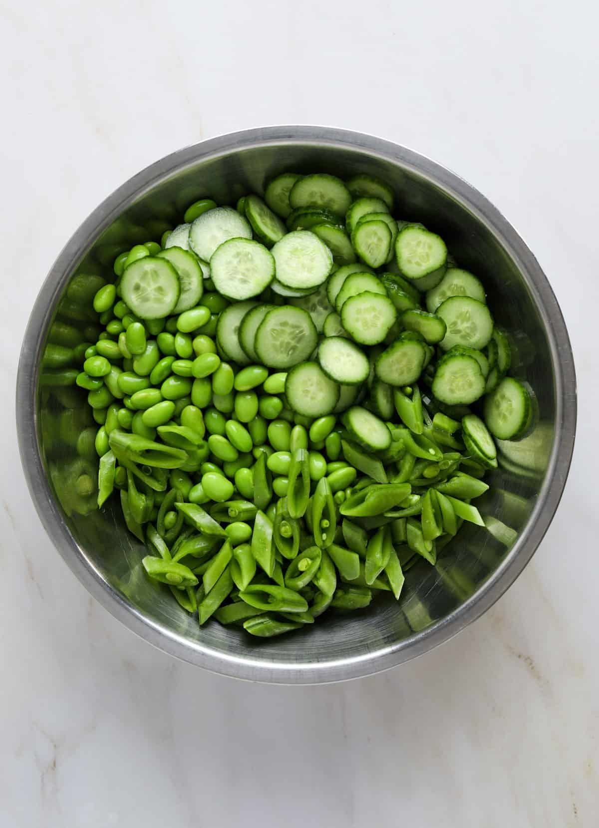 a stainless steel mixing bowl filled with edamame, sliced cucumbers and chopped snap peas.