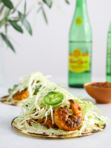 A close up image of a taco with shrimp and cabbage. Two green bottles of Topo Chico mineral water in the background.