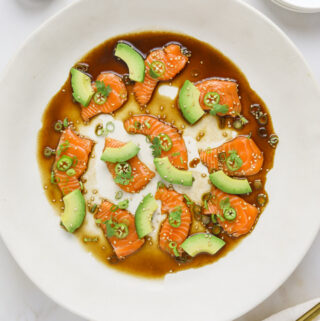 a white plate with slices of raw salmon, avocado, soy sauce, sesame seeds with chop sticks on the side.