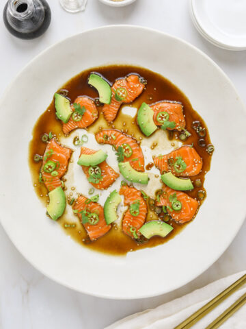 a white plate with slices of raw salmon, avocado, soy sauce, sesame seeds with chop sticks on the side.