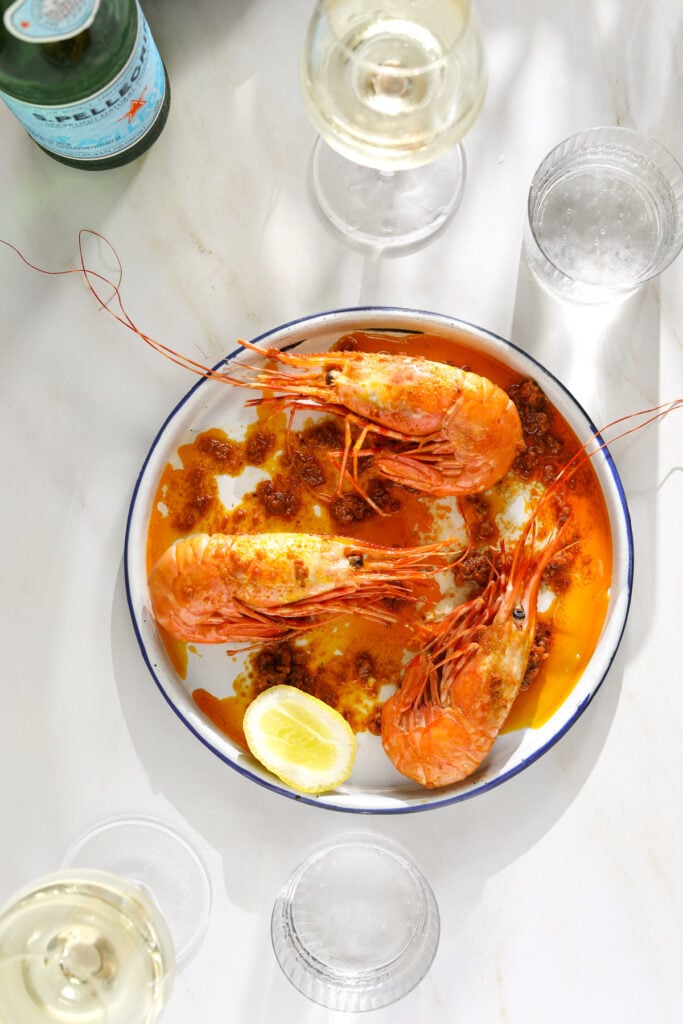 An enamel white plate with three cooked prawns topped with red sauce and a slice of lemon.
