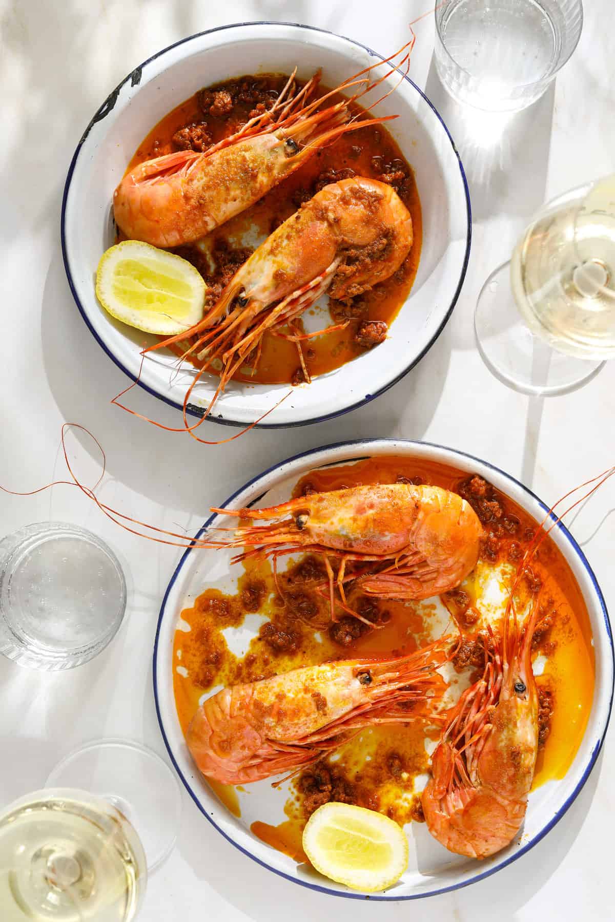 Two bowls of whole , cooked prawns with a red sauce, lemon wedges and wine glasses on the side.