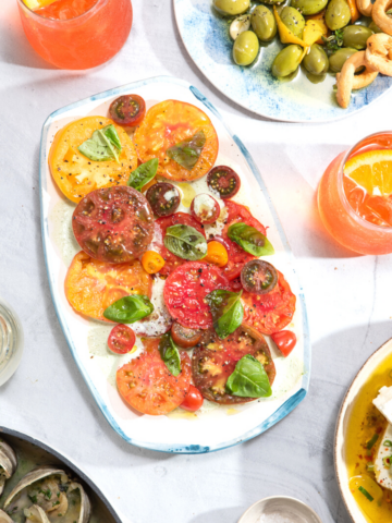A vibrant red tomato salad with fresh basil on a rectangular platter surrounded by cocktails on ice with orange wedges.