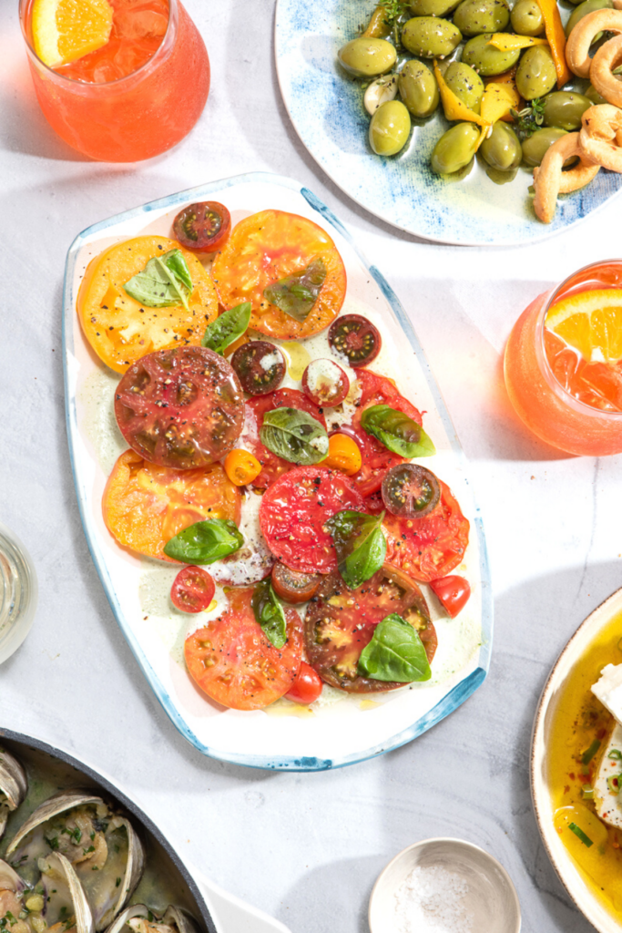 A vibrant red tomato salad with fresh basil on a rectangular platter surrounded by cocktails on ice with orange wedges.