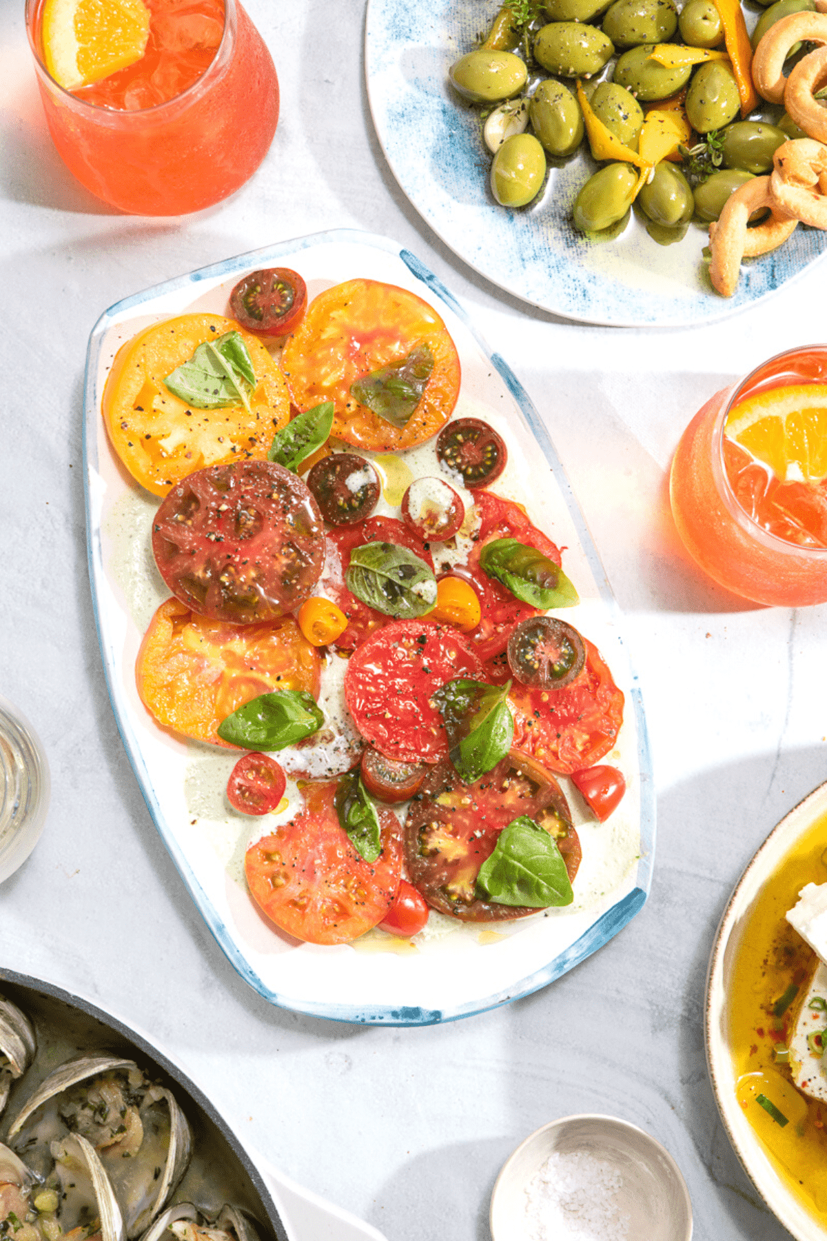 A vibrant red tomato salad with fresh basil on a rectangular platter surrounded by cocktails on ice with orange wedges.