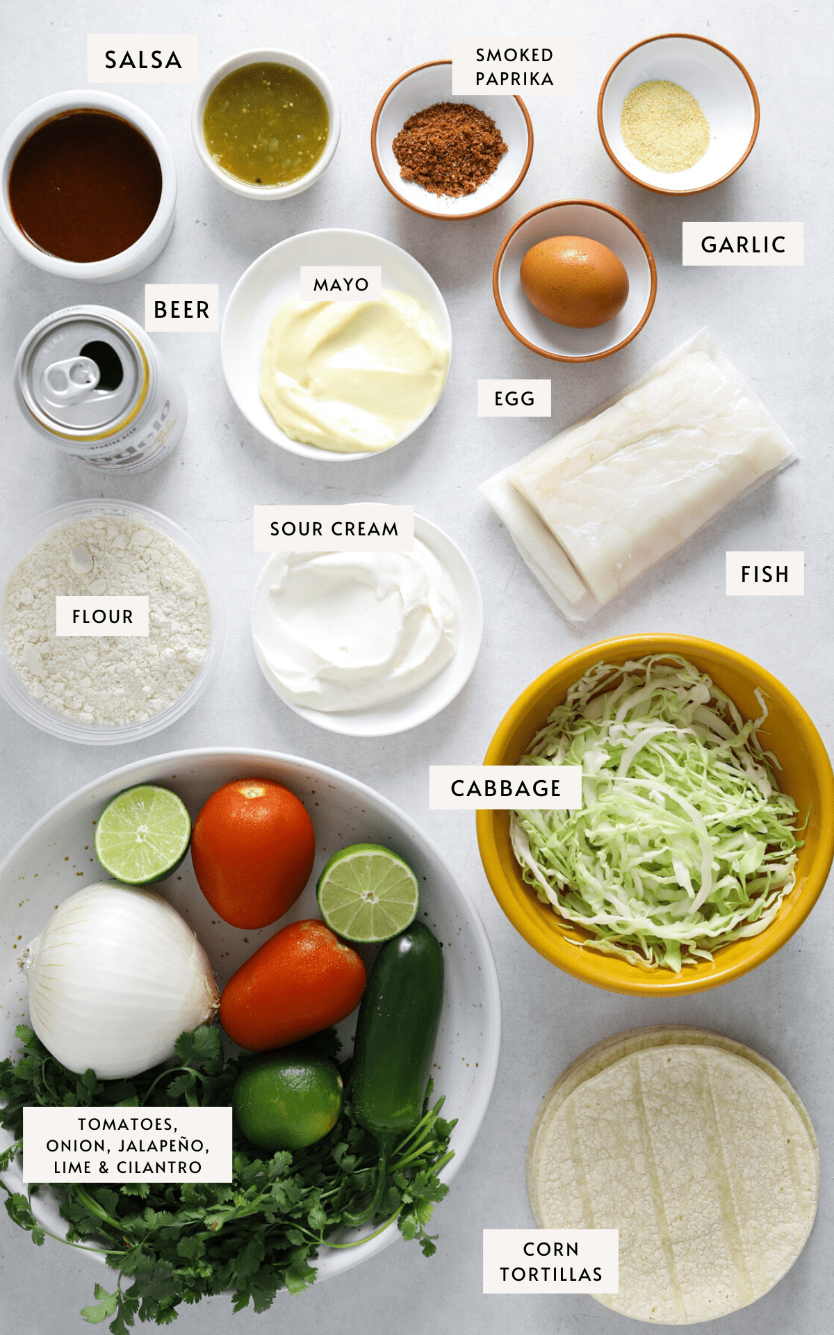 Recipe ingredients portioned into individual bowls; shredded cabbage, a fish filet, flour, sour cream, salsa, limes, a stack of corn tortillas.
