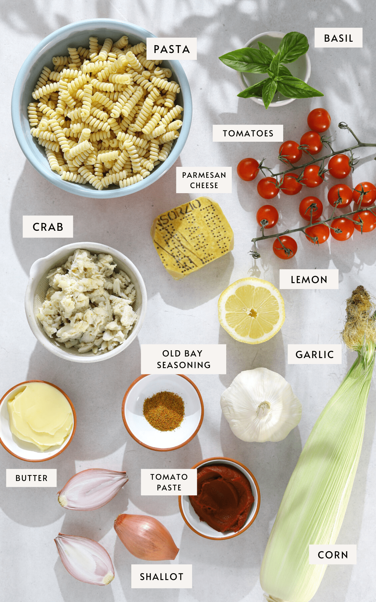 Recipe ingredients portioned into small bowls; picked crab meat, short-cut pasta, basil leaves, cherry tomatoes, paprika, tomato paste, halved shallots and a whole ear of corn.