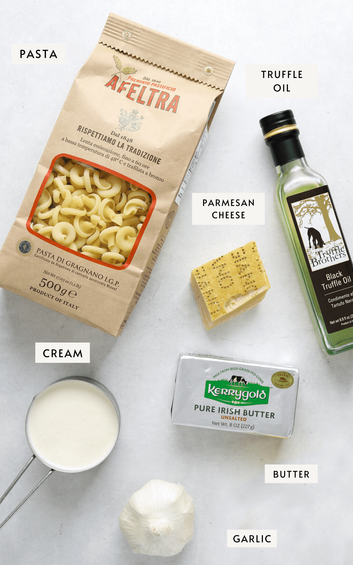 A brown paper bag of pasta, a bottle of black truffle oil, a pack of butter, a hunk of parmesan cheese, a measuring cup filled with cream, one lemon and a head of garlic all on a light blue-gray background.