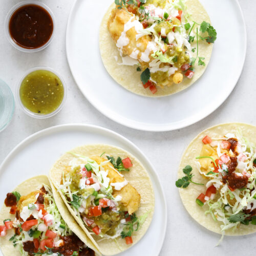 Two small white plates filled with tacos and surrounded by cups of red and green salsa and cans of Mexican beer.