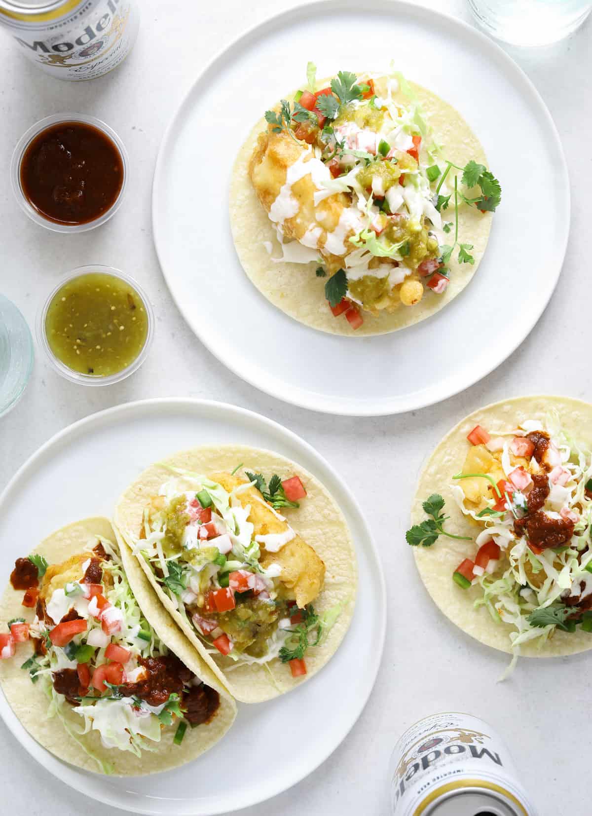 Two small white plates filled with tacos and surrounded by cups of red and green salsa and cans of Mexican beer.