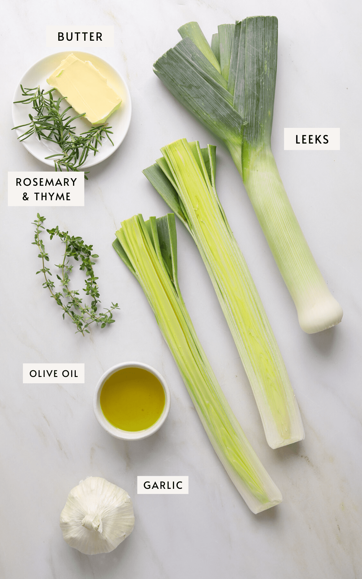 Two leeks (one whole, one cut in halt lengthwise), a small bowl of olive oil, a knob of butter, a head of garlic and sprigs of rosemary and thyme on a white marble background.