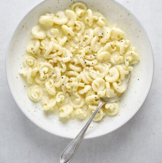 A white bowl of pasta on a blue-grey background with a silver spoon.