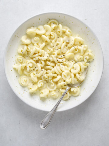 A white bowl of pasta on a blue-grey background with a silver spoon.