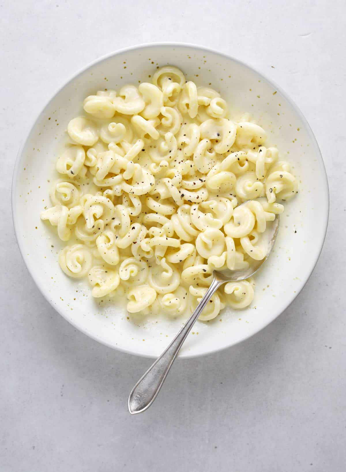 A white bowl of pasta on a blue-grey background with a silver spoon.