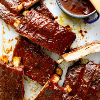 A baking tray with barbecue ribs cut into pieces and a small dish of sauce in a blue bowl.