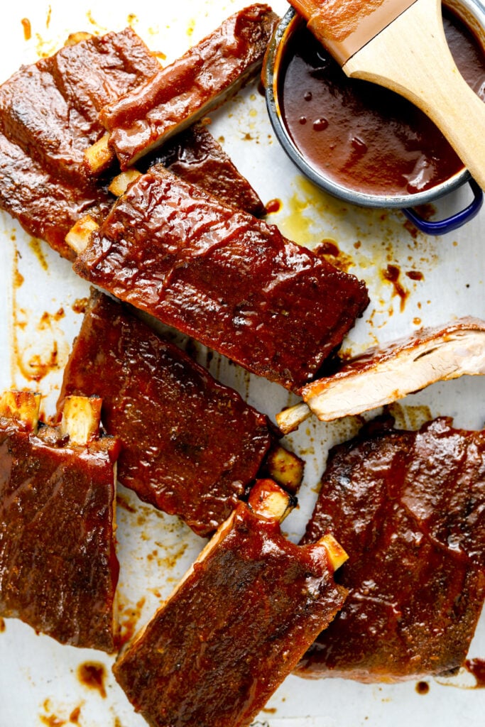 A baking tray with barbecue ribs cut into pieces and a small dish of sauce in a blue bowl.