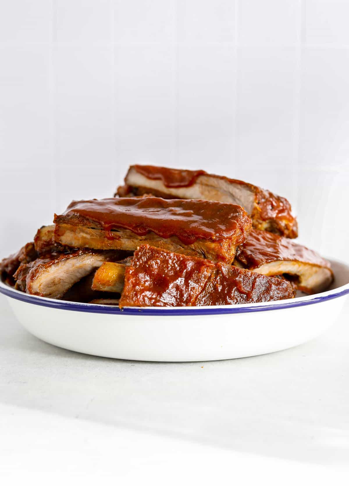 A white enamel bowl with a blue rim filled with BBQ spare ribs slathered with barbecue sauce.