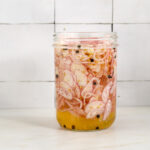 A mason jar filled with thinly sliced pickled shallots in bring with black peppercorns and mustard seeds.