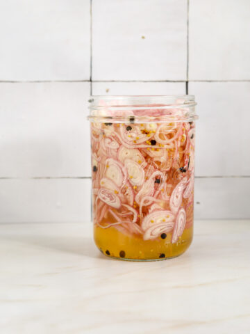 A mason jar filled with thinly sliced pickled shallots in bring with black peppercorns and mustard seeds.