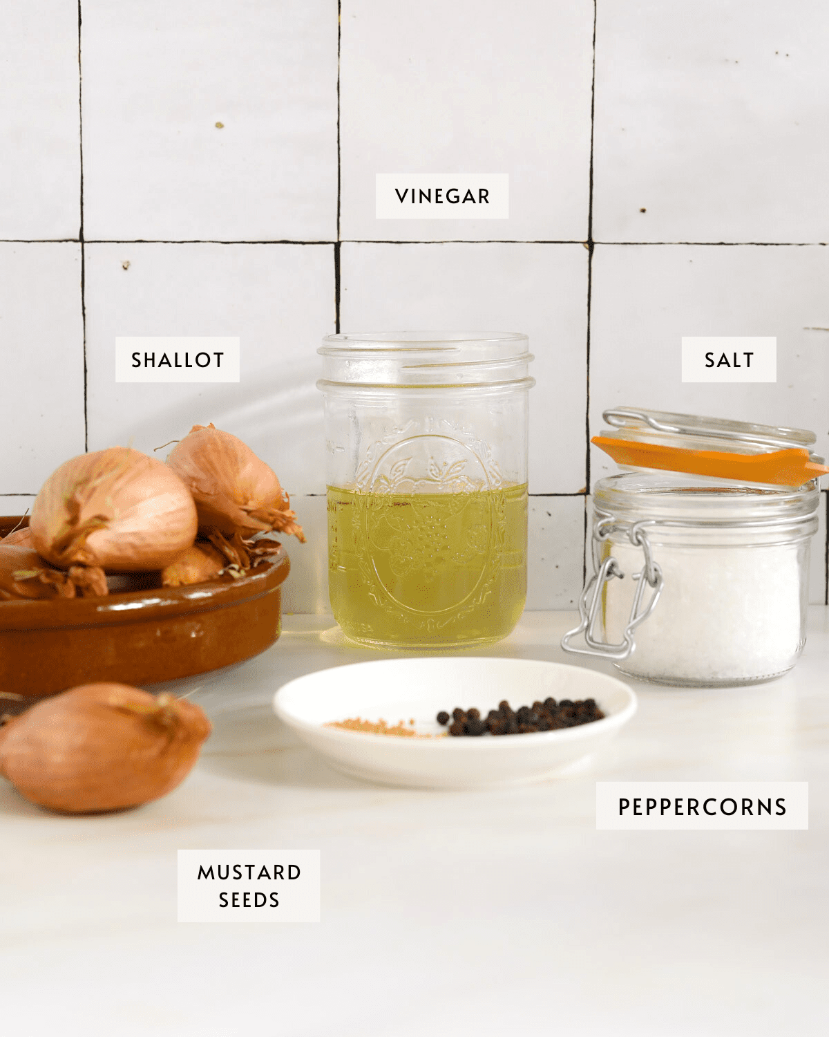 A small bowl of whole shallots, a jar of vinegar, a small bowl of pepper and mustard seeds, a glass jar of kosher salt.