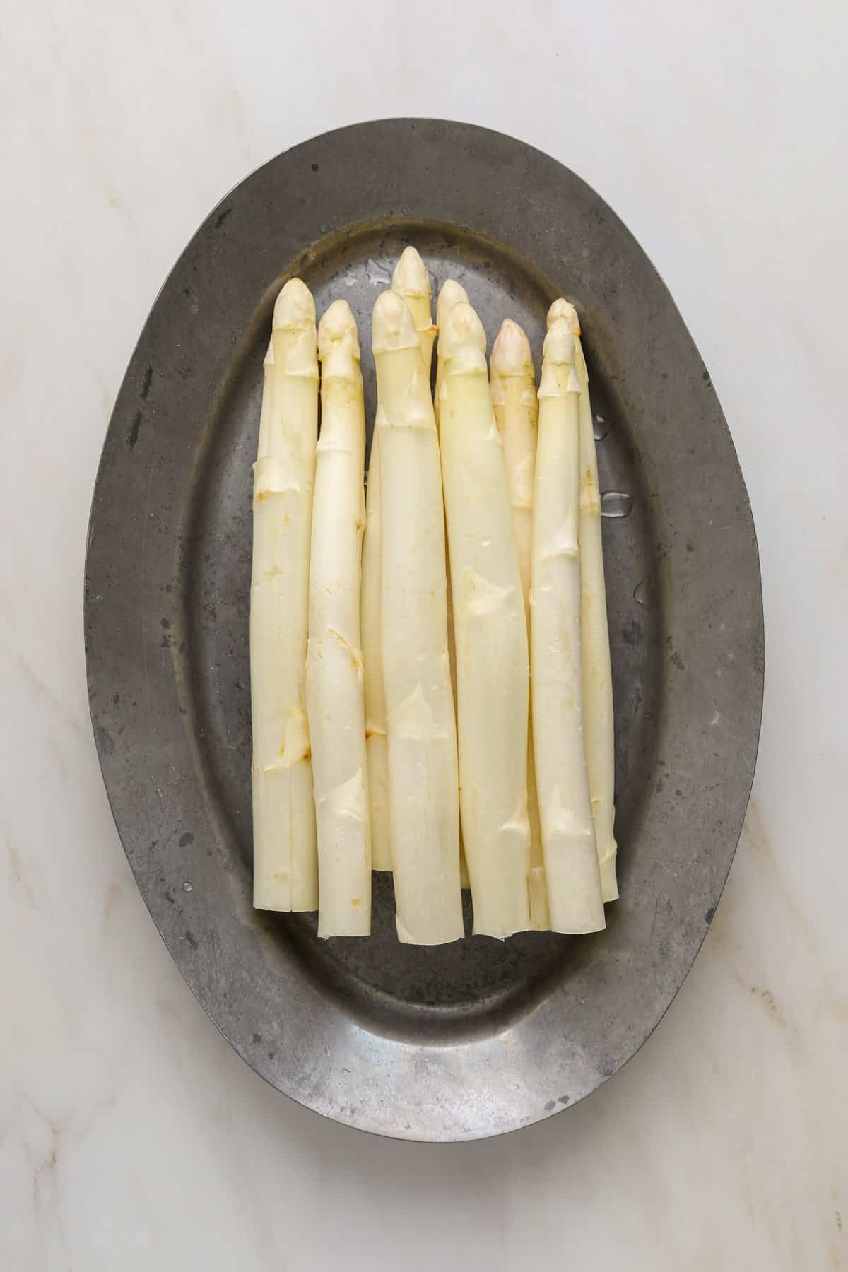 White asparagus spears on a silver platter resting on a marble stone table top.