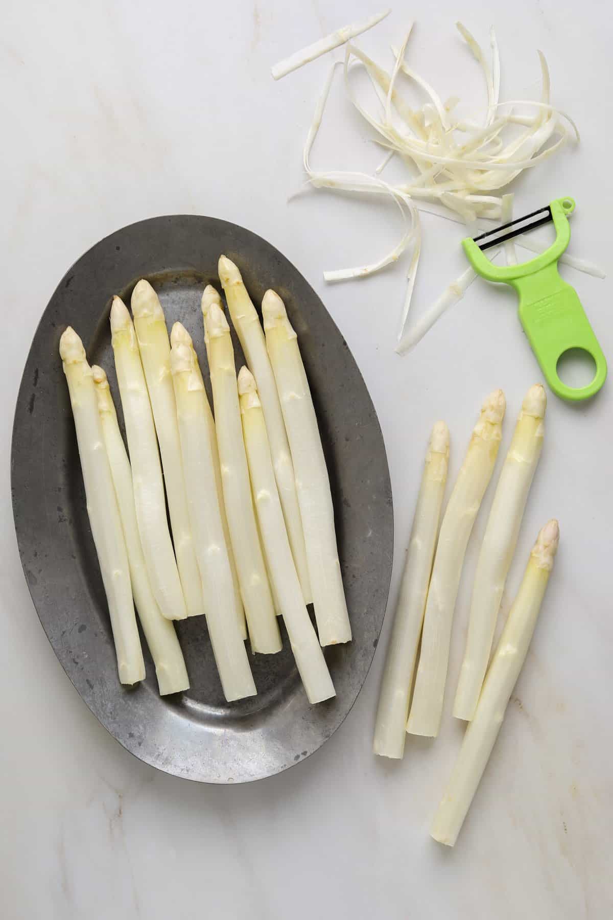 A metal oval platter with peeled white asparagus with a vegetable peeler on side and a small pile of asparagus shavings.