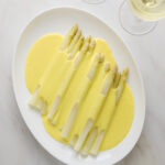 A white oval platter with white asparagus spears covered in hollandaise sauce with two glasses of white wine on the side.