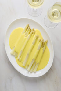 A white oval platter with white asparagus spears covered in hollandaise sauce with two glasses of white wine on the side.