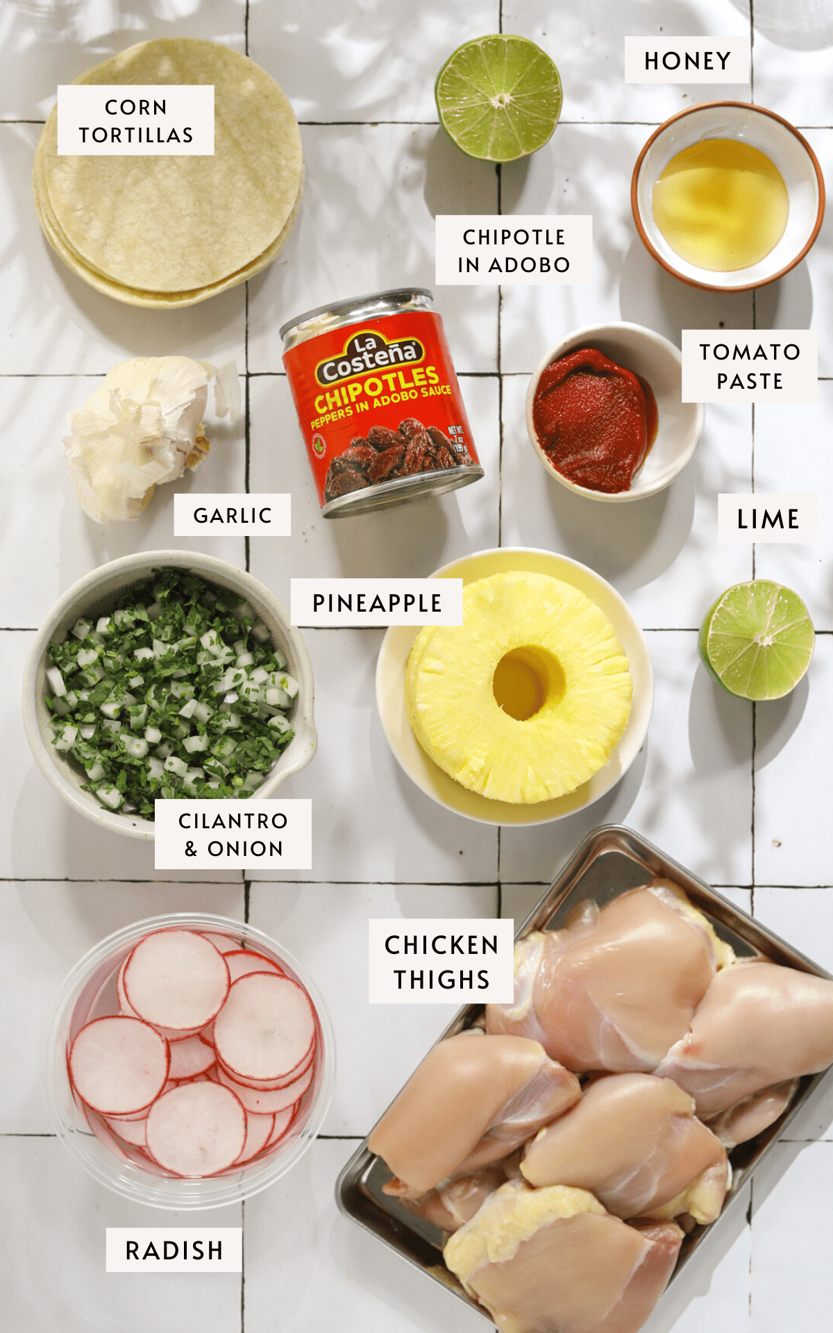 A stack of corn tortillas, small bowls of ingredients like; tomato paste, honey, sliced radish, chopped cilantro and onion. A small white bowl of pineapple, a can of chipotle peppers and a tray of raw chicken.