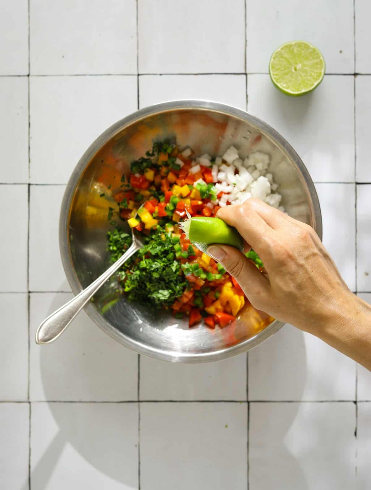 A mixing bowl filled with chopped salsa ingredients and a hand squeezing lime juice into the bowl.