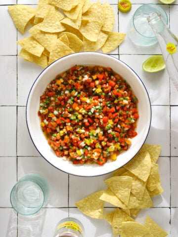 A white bowl of vibrant red and yellow pico de gallo salsa on a tile background surrounded by tortilla chips.