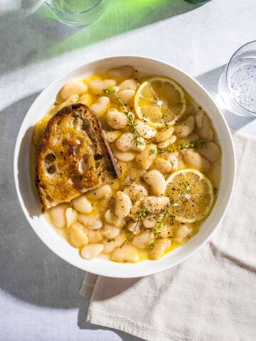 A white bowl filled with beans, lemon slices, fresh thyme stems and a thick slice of golden brown toasted bread.