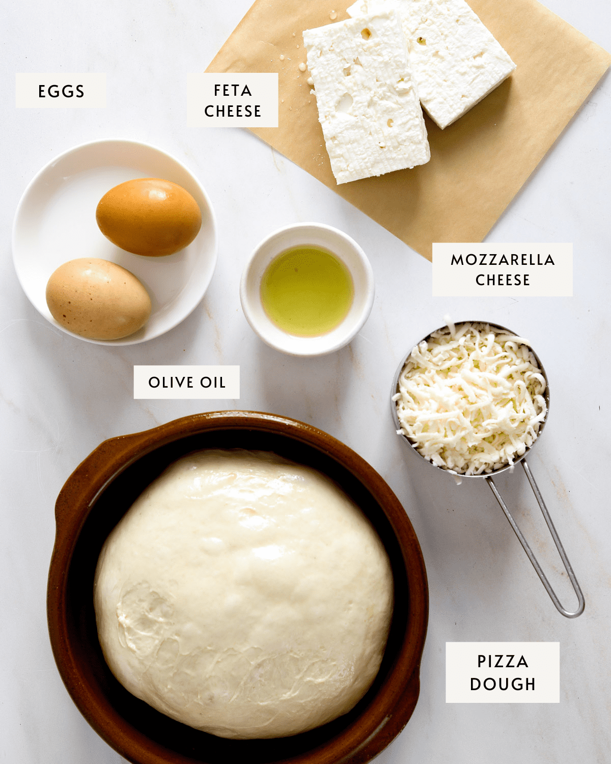 A small white bowl with two brown eggs, two hunks of feta cheese, a ball of pizza dough in a ceramic bowl, a measuring cup with grated mozzarella cheese and a small dish of olive oil.