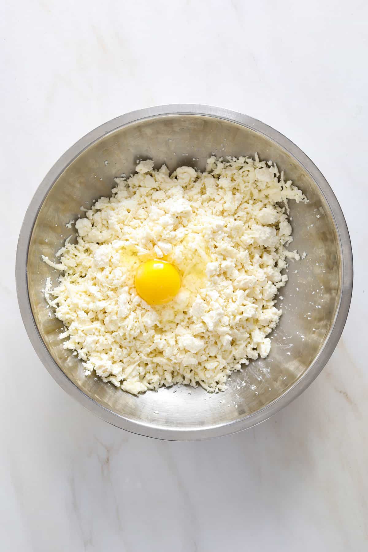 A stainless steel mixing bowl filled grated cheese and a whole egg.