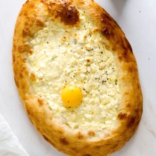 An oval "Khachapuri Pizza" baked into an oval shape with golden brown crust and filled white melty cheese sprinkled with cracked black pepper and topped with an egg yolk.
