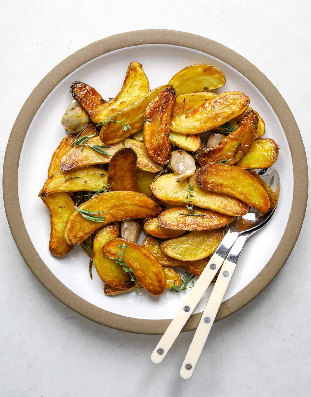 A round white platter filled with roasted fingerling potatoes and two white and silver serving spoons off to the side.