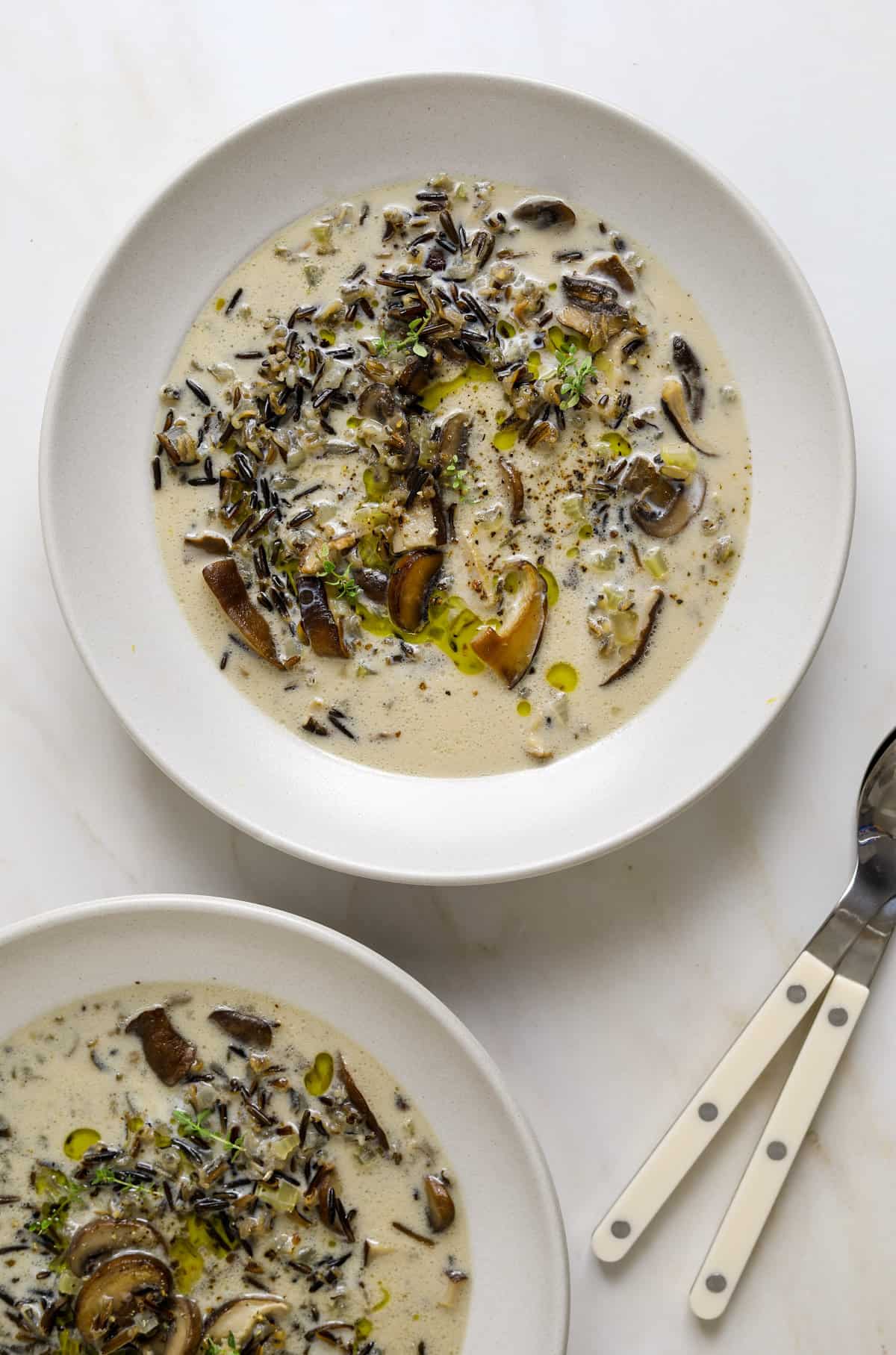 A white bowl filled wild rice soup with a drizzle of olive oil and fresh thyme leaves on a white background.