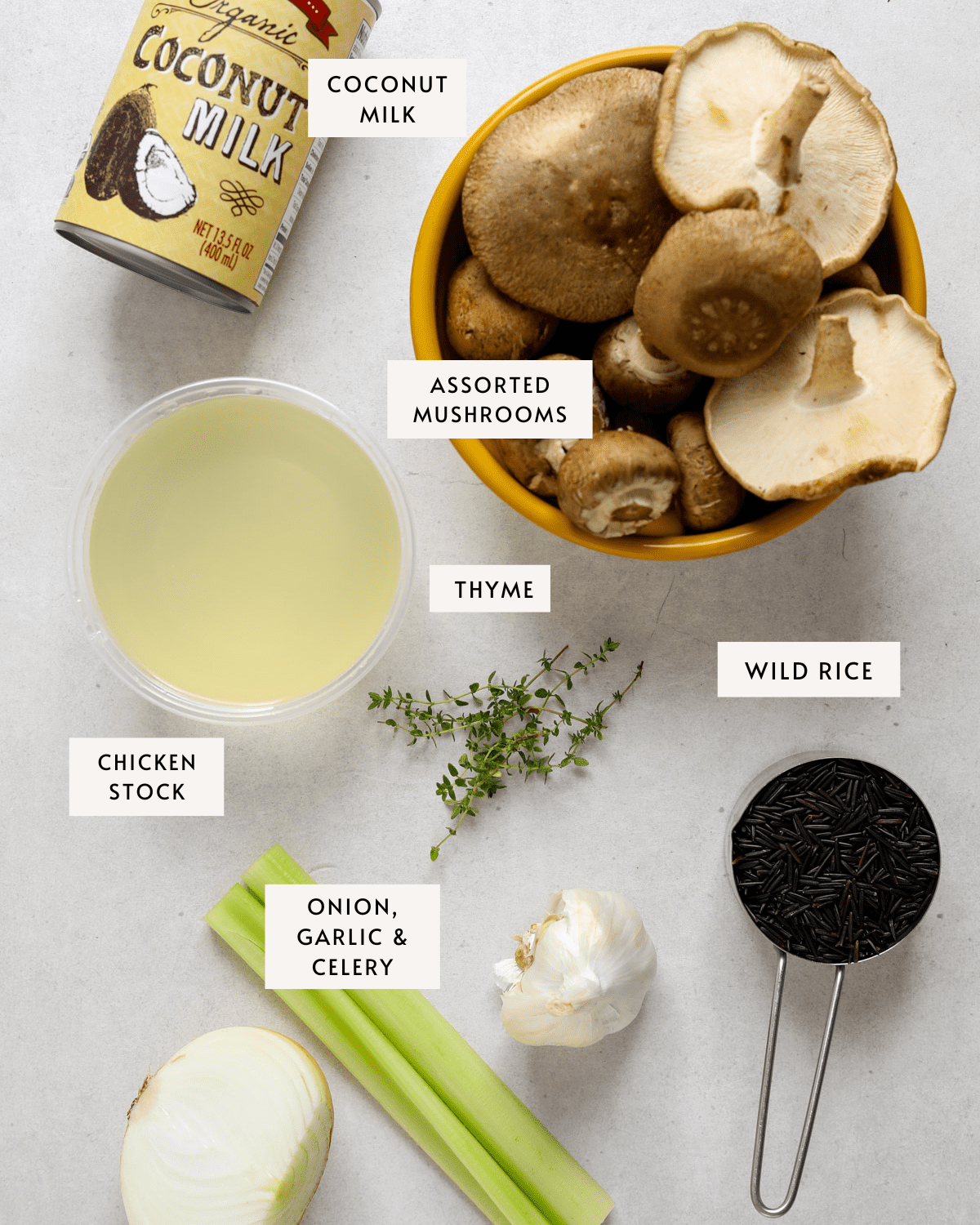 A can of coconut milk, a small plastic container of chicken stock, a measuring cup with wild rice, a yellow bowl of assorted mushrooms, half and onion a head of garlic and a bundle of fresh thyme stems.