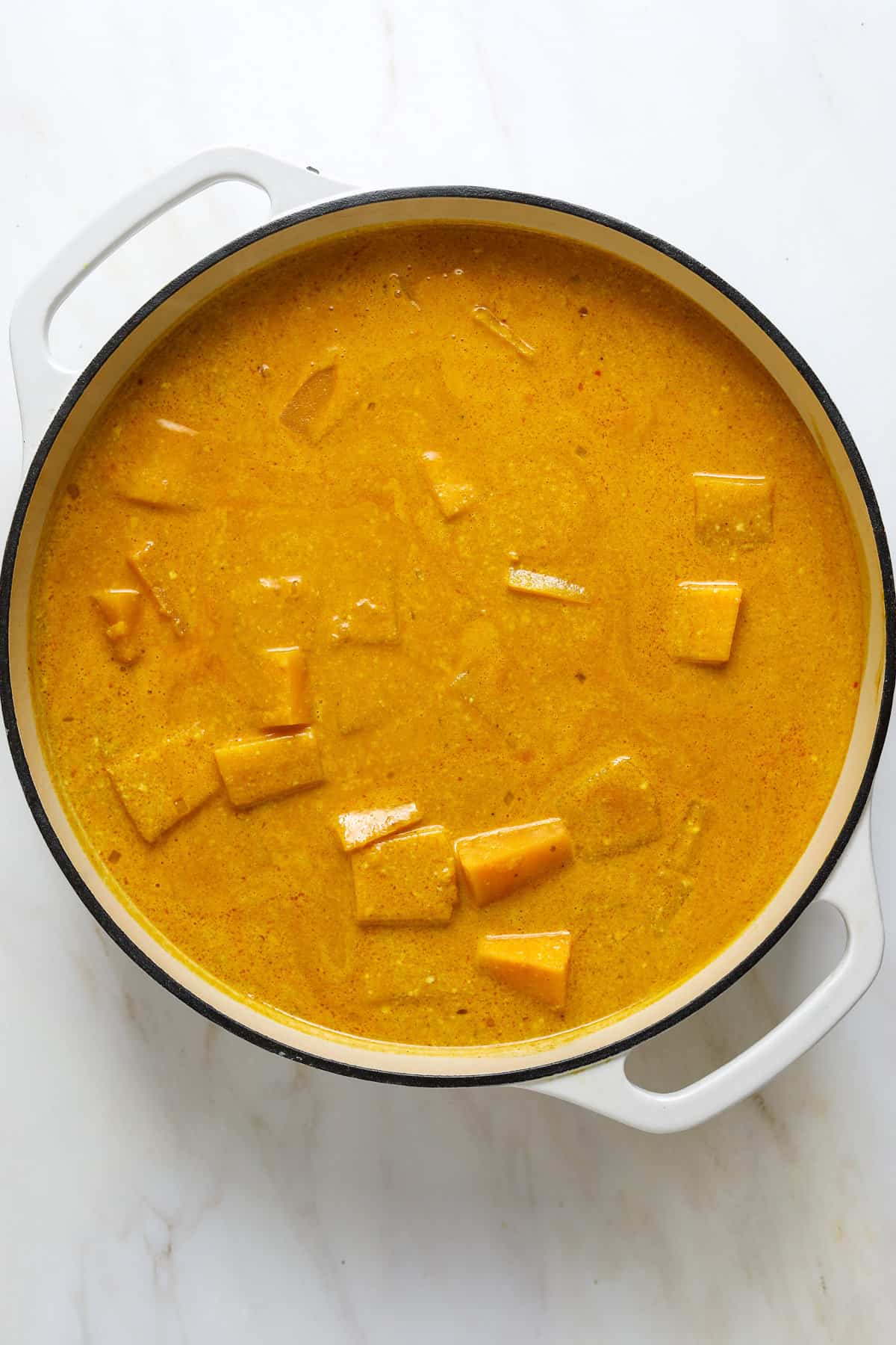 A large round, white enamel sauté pan brimming with pale orange curry sauce and chunks of butternut squash.