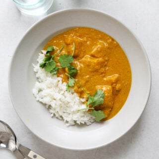 Overhead view of a white bowl of curry and rice topped with fresh cilantro stems. Two spoons on the side as well as a glass of water and a bottle of sparkling water.