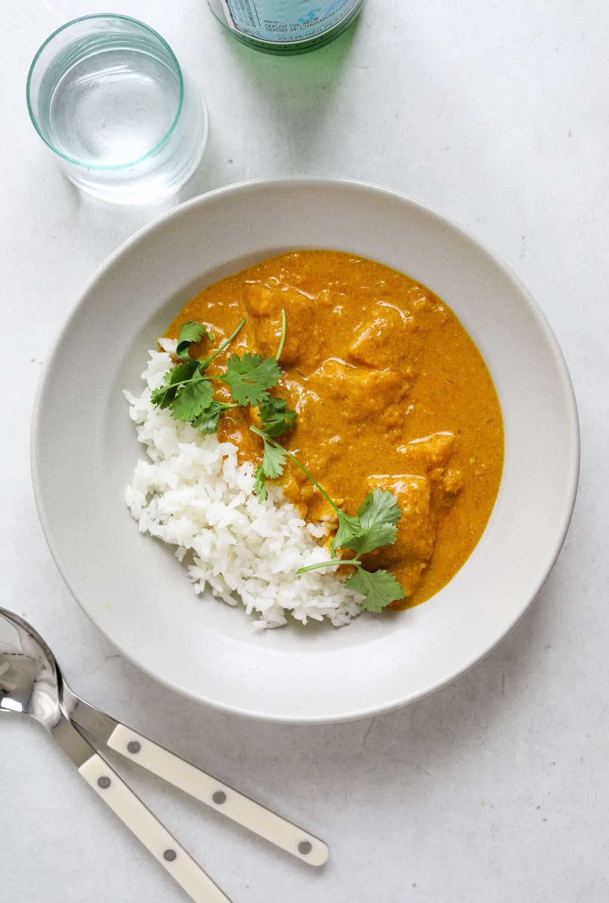 Overhead view of a white bowl of curry and rice topped with fresh cilantro stems. Two spoons on the side as well as a glass of water and a bottle of sparkling water.