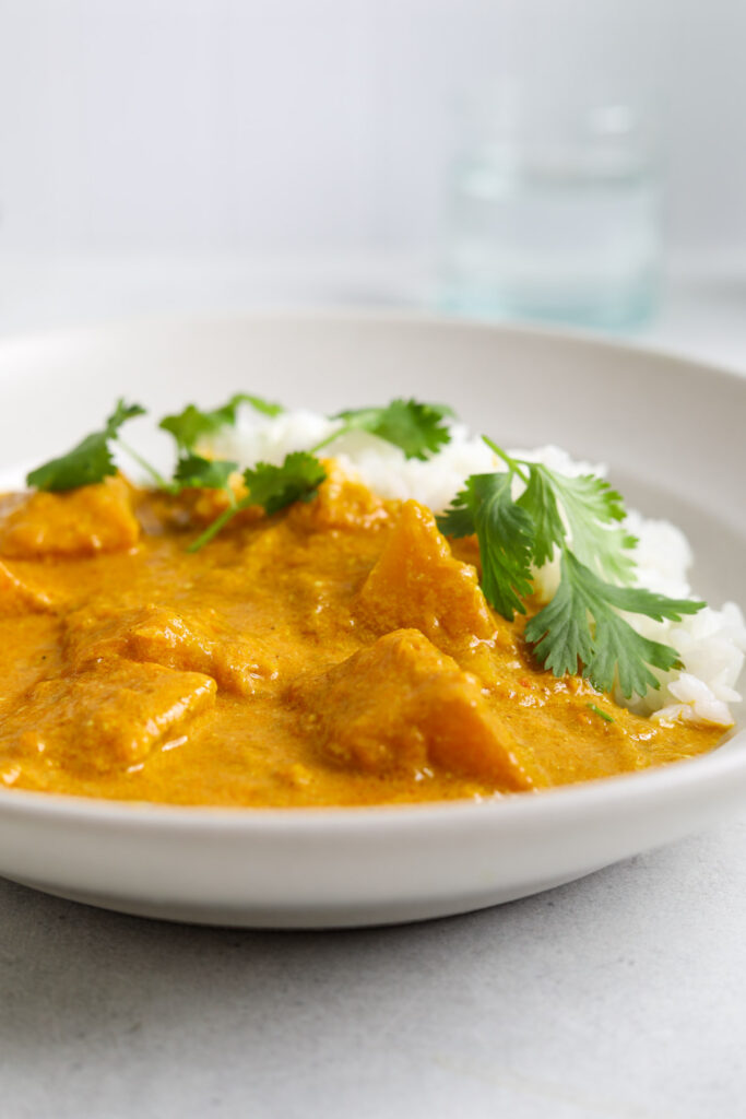 A side profile of a white bowl filled with white rice, pale orange butternut squash curry with a few pieces of fresh cilantro on top.